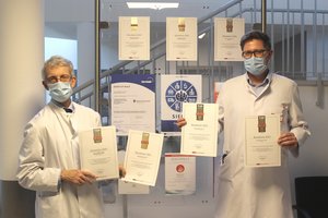 Die Chefärzte der Klinik stehen vor der Zertifikatswand des Krankenhauses und halten die neuen Zertifikate hoch.